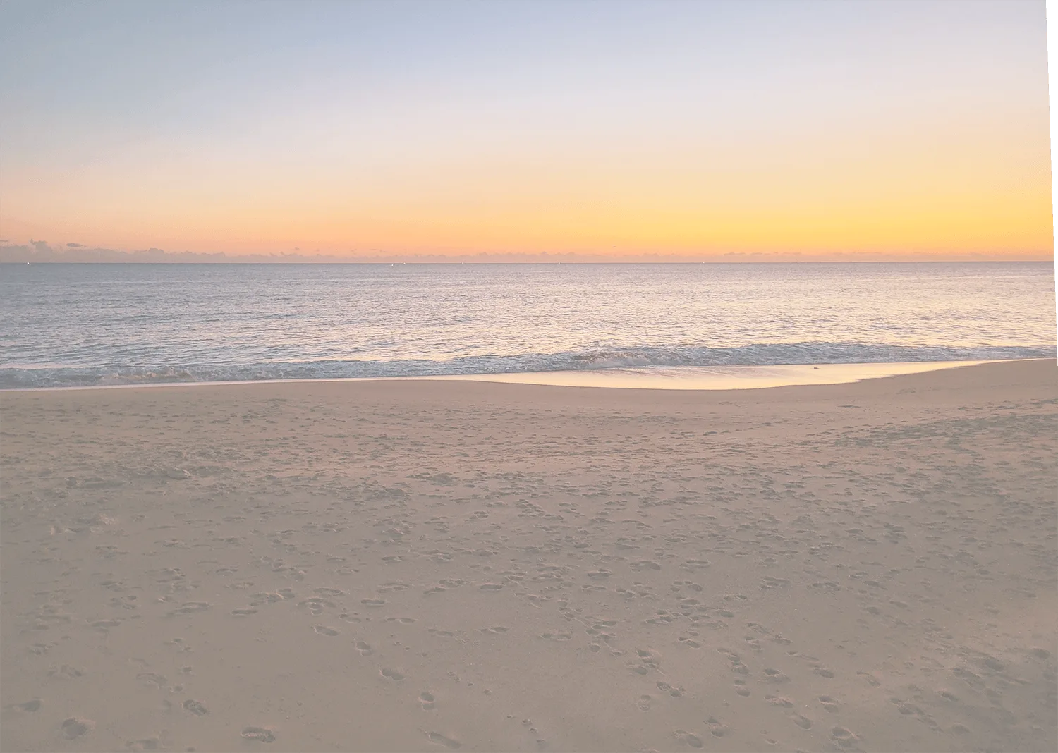 Sunset over a calm sea with a sandy beach and footprints scattered across the shore.